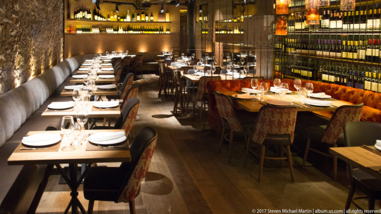 Long restaurant dining room with set tables, upholstered banquettes, warm lighting, and floor-to-ceiling wine shelving.