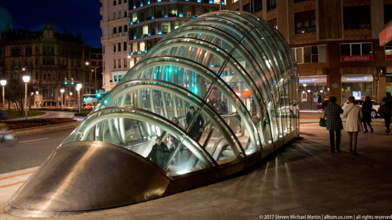 Curved transparent glass-and-steel Bilbao Metro entrance canopy glowing at night beside city buildings and pedestrians.