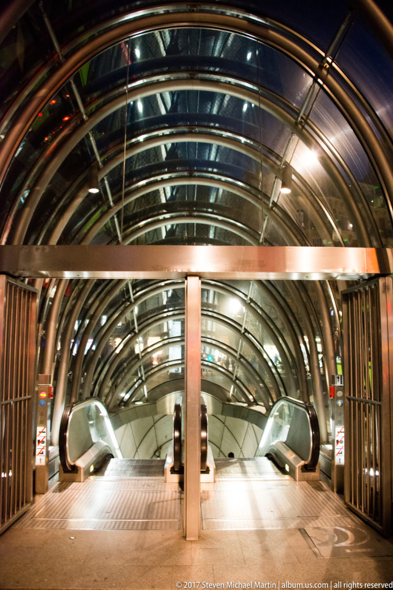 Paired escalators descend beneath a curved ribbed glass canopy at a Bilbao Metro entrance illuminated at night.