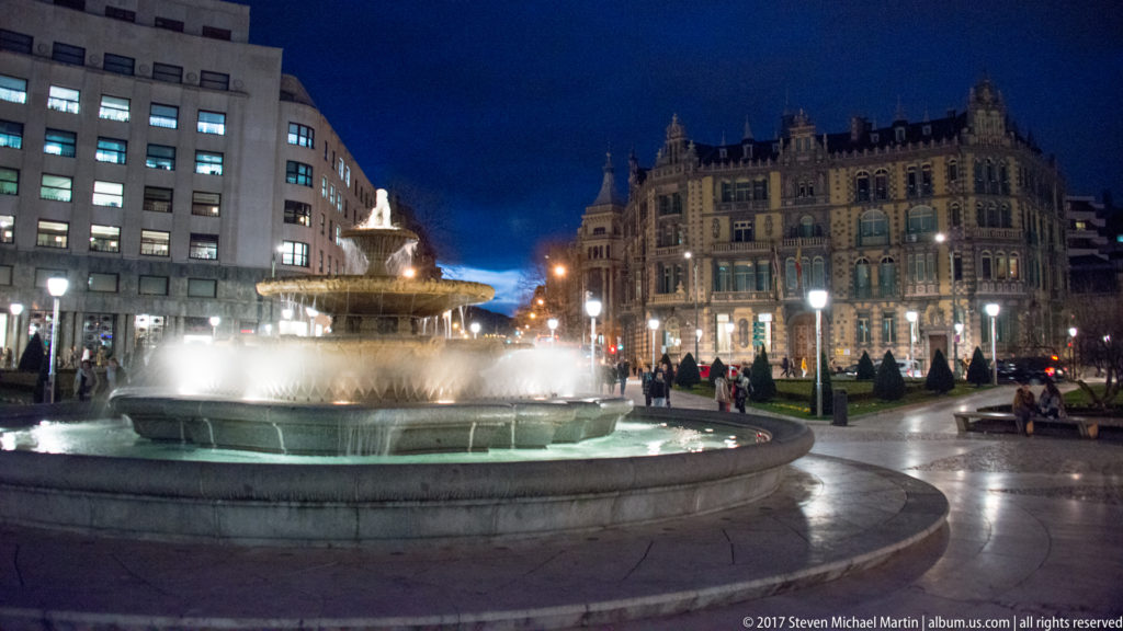 Large circular fountain illuminated at night, with cascading spray, pedestrians, and ornate central Bilbao buildings behind it.