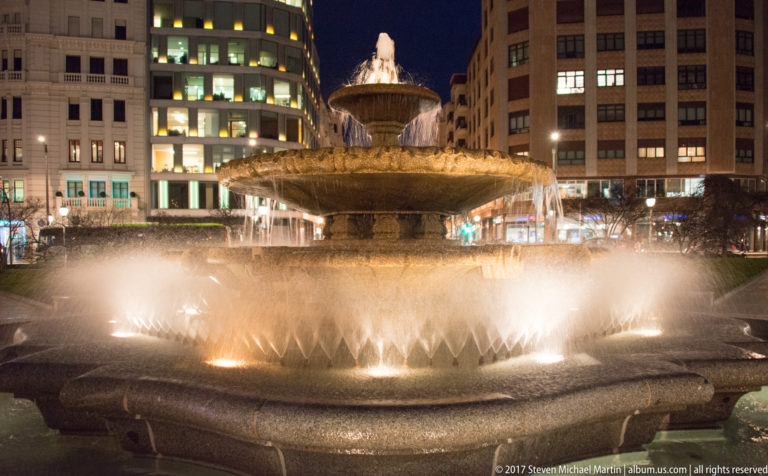 Close nighttime view of a tiered stone fountain with illuminated jets and water cascading into a broad circular basin.