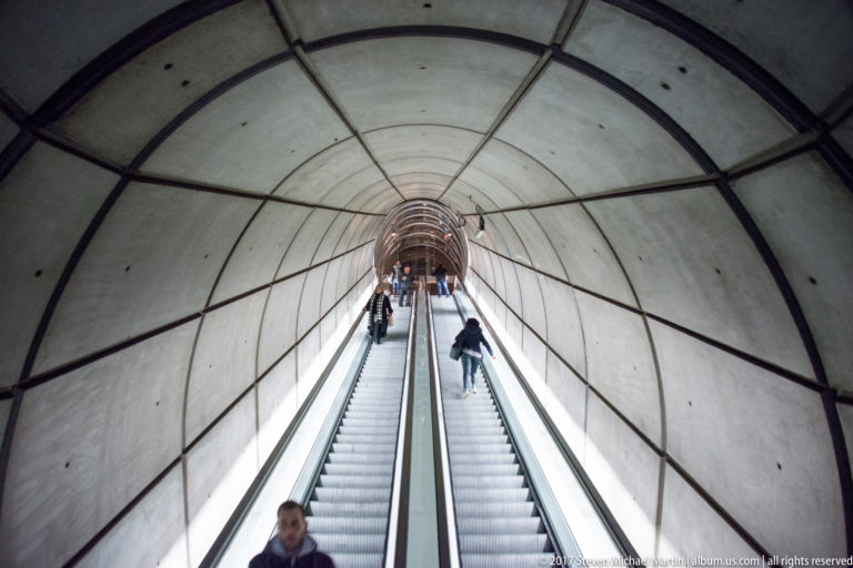 Long escalator rises through a curved concrete Bilbao Metro passage with several riders spaced along the incline.