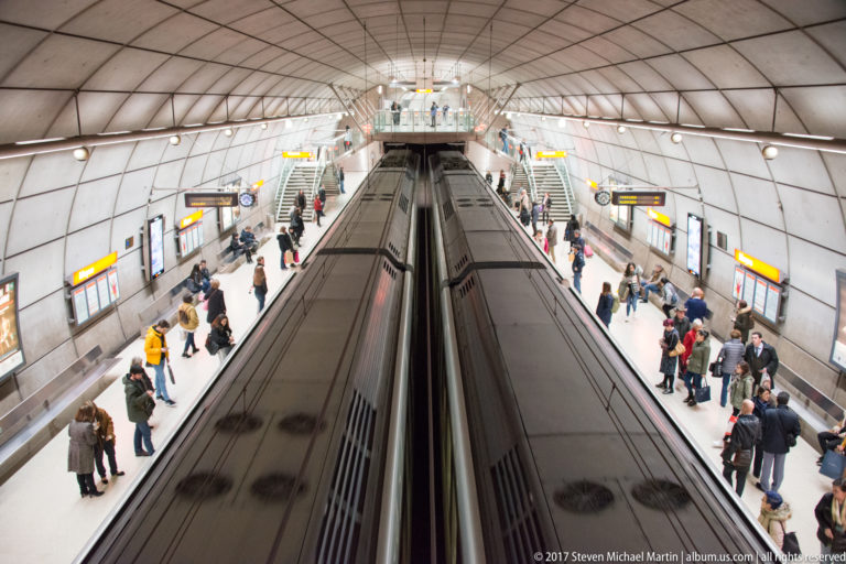 Metro train between two busy platforms beneath a broad curved ceiling in a Bilbao Metro station.