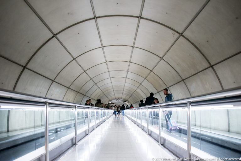 Long moving walkway with glass rails inside a bright curved tunnel, carrying pedestrians through a Bilbao transit passage.