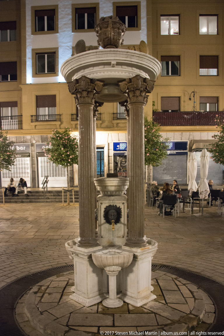 Small stone fountain with fluted columns, carved capitals, circular canopy, and central basin in a Bilbao square at night.