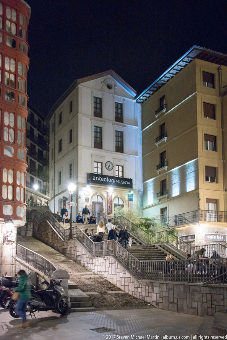 Bizkaia Archaeological Museum at night with “arkeologi museoa” sign, broad steps, railings, and café seating below.