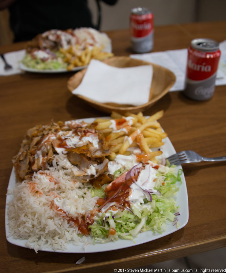 Rectangular plate piled with sliced roasted meat, rice, fries, lettuce, tomato, and sauce on a restaurant table.