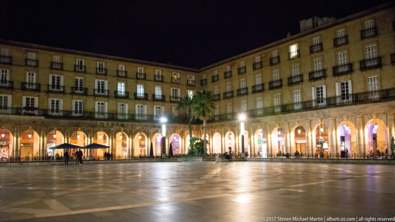 Night view across Bilbao’s Plaza Nueva with continuous illuminated arcades, uniform façades, reflective paving, and a few palm trees.