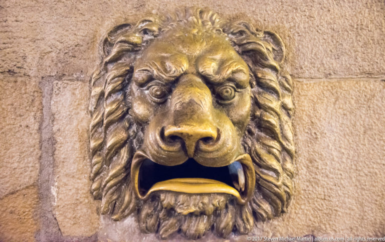 Close frontal view of a bronze lion head with deeply modeled mane and open mouth mounted on a rusticated stone wall.