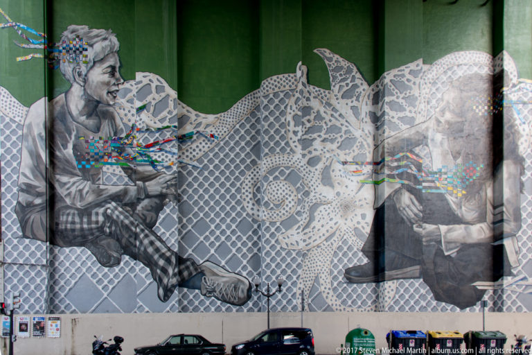 Large black-and-white mural of two seated figures overlaid with lace-like patterns and small bands of color.