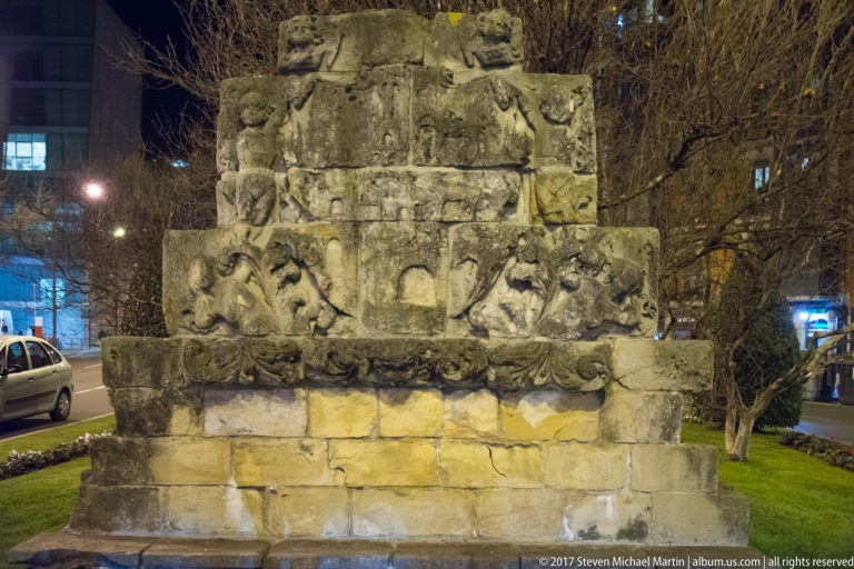Weathered stone monument at night with worn figurative and heraldic reliefs on stepped blocks above a plain masonry base.