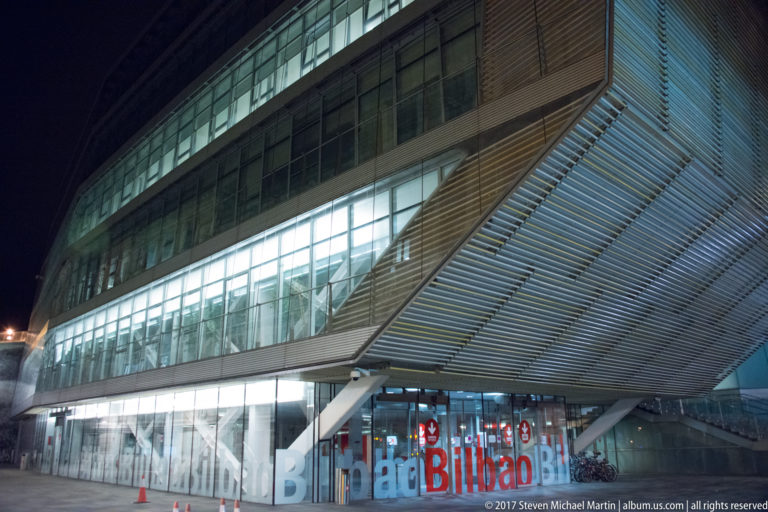 Modern Bilbao building with horizontal glass bands, a large projecting metal louvered canopy, interior stairs, and oversized street-level lettering.