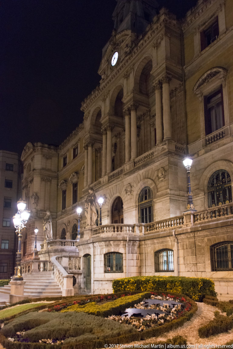 Illuminated Bilbao City Hall with tall central columns, arched loggia, clock, stone balustrades, and formal garden beds.