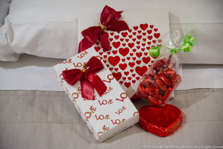 Valentine gifts on white bedding, including heart-patterned boxes, red bows, a heart-shaped chocolate, and a bag of red sweets.