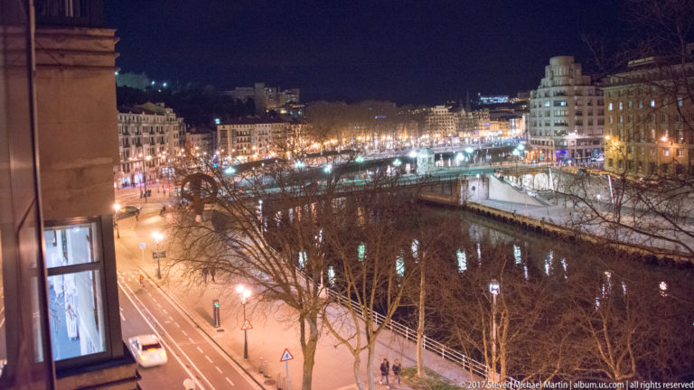 Elevated nighttime view of Bilbao’s riverfront with dark water, illuminated quays, traffic, buildings, and bare trees in the foreground.