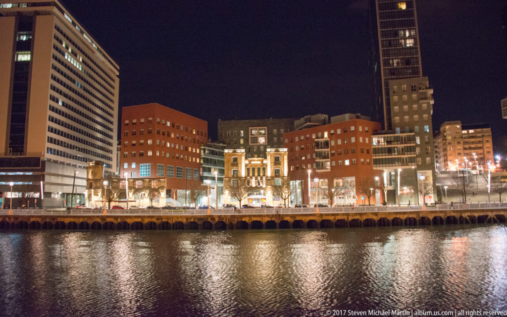 Modern Bilbao towers and office blocks illuminated across the Nervión estuary, with long reflections shimmering on the water.