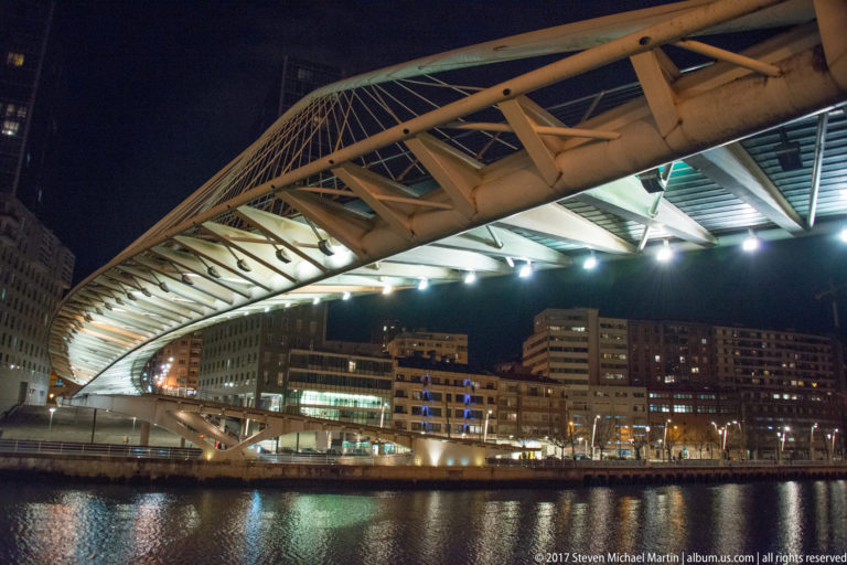 Illuminated white Zubizuri pedestrian bridge curves over the Nervión at night, with cable lattice and city buildings beyond.