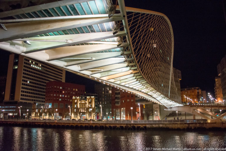 Close view beneath Zubizuri showing white steel ribs, translucent deck panels, cable netting, and illuminated buildings across the river.