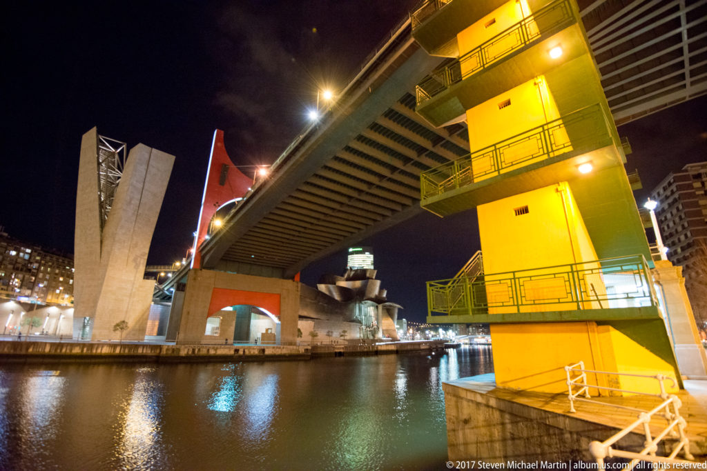 Night view beneath a Bilbao road bridge with a bright yellow support pier, dark deck, red structural element, and reflections on the river.