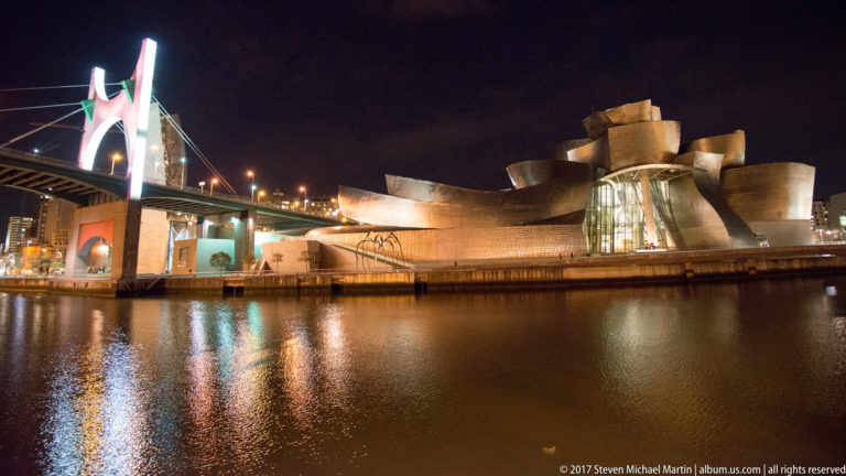 Night view across the Nervión to the Guggenheim Museum Bilbao beside the red arch and deck of La Salve Bridge.