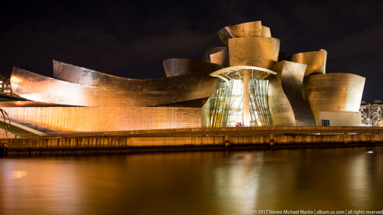 Guggenheim Museum Bilbao at night, with curving titanium volumes and a tall illuminated glass atrium reflected in the river.