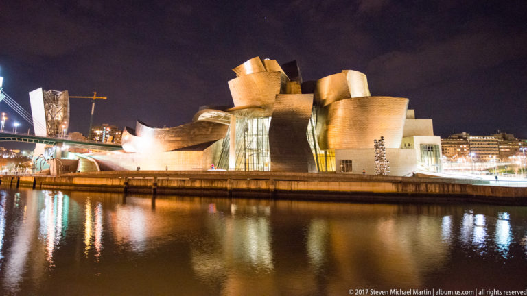Wide night view of the Guggenheim Museum Bilbao beside La Salve Bridge, with bright reflections across the Nervión.