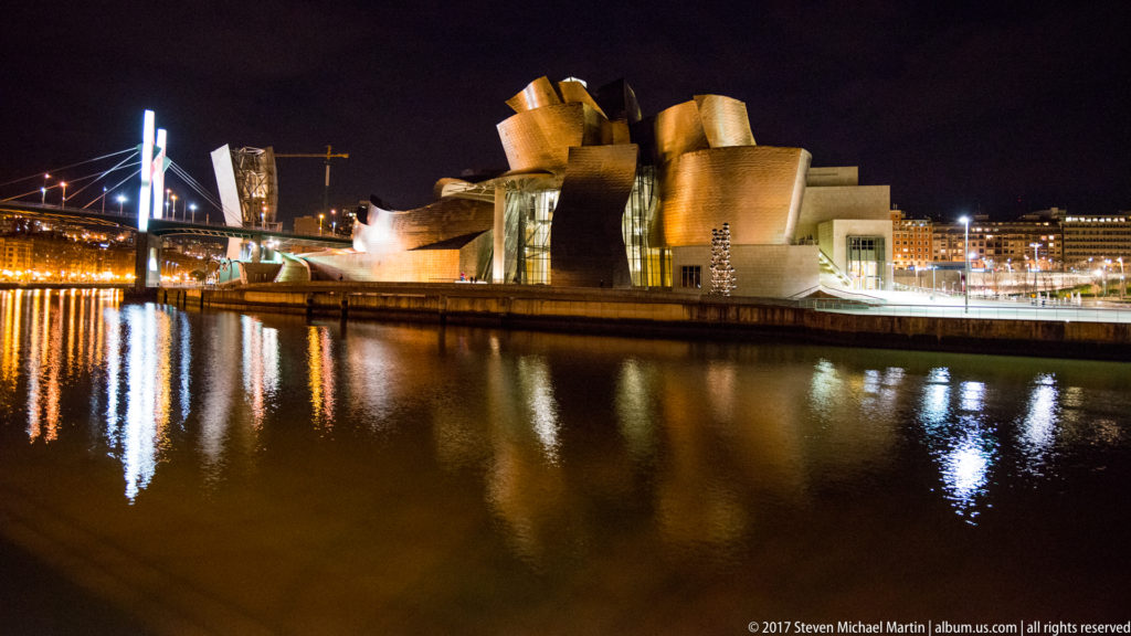 Guggenheim Museum Bilbao illuminated across the Nervión, with broad dark water and vertical reflections in the foreground.