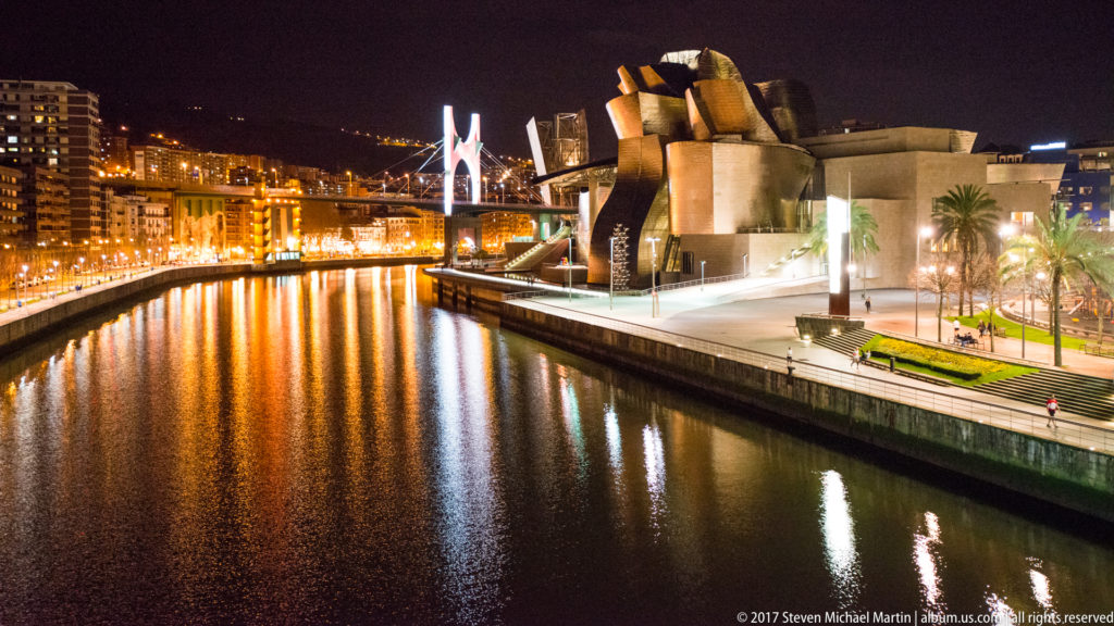 Night view along the Nervión toward the Guggenheim Museum Bilbao and La Salve Bridge, with illuminated quays and hillside buildings.