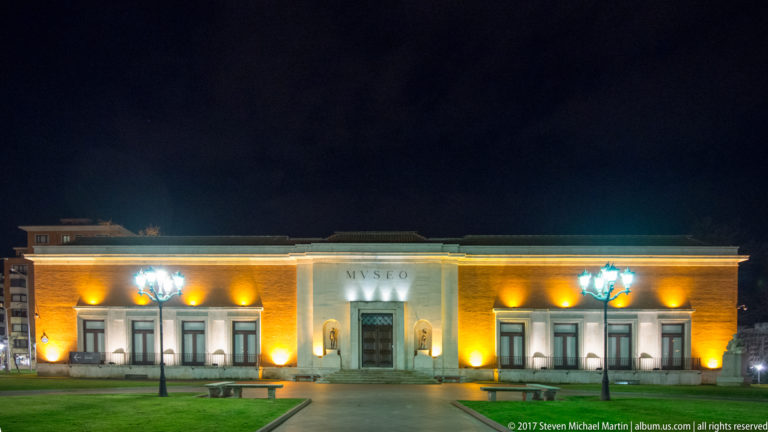 Bilbao Fine Arts Museum at night, with a low symmetrical façade, central “MUSEO” inscription, lawn, and illuminated entrance.