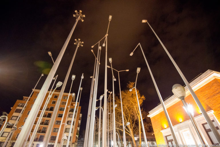 Tall white streetlight poles lean at varied angles against the night sky, framed by Bilbao buildings at the edges.
