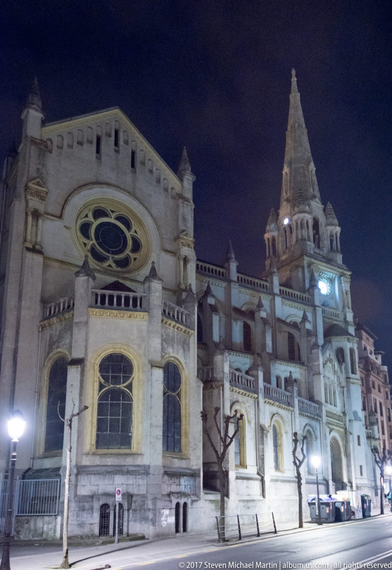 Historic Bilbao church at night with a large rose window, tall illuminated bell tower, arched windows, and bare street trees.