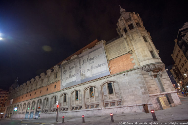 Long brick-and-stone Azkuna Zentroa façade at night with faded historical lettering, repeated arches, and an ornate corner tower.