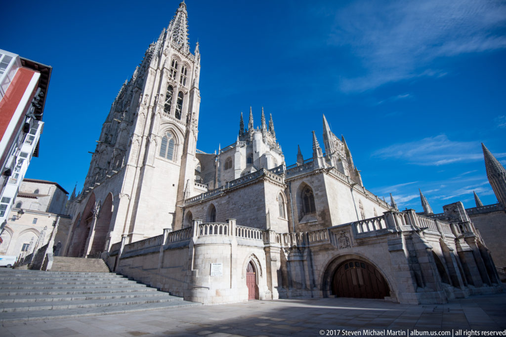 Wide exterior view of Burgos Cathedral from a paved plaza, with tall Gothic tower, chapels, pinnacles, and blue sky.