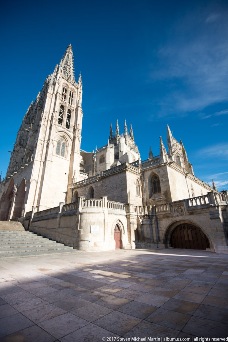 Burgos Cathedral side elevation with bell tower, lower chapels, arched entrances, pinnacles, and broad paved plaza.