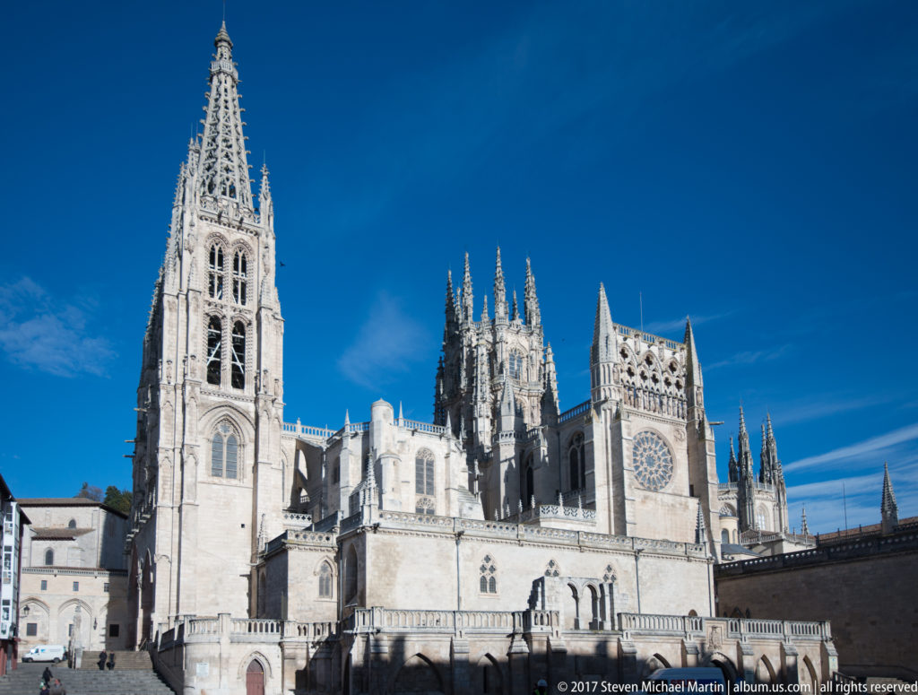 Wide view of Burgos Cathedral showing a tall Gothic tower, clustered pinnacles, buttresses, and a large rose window.