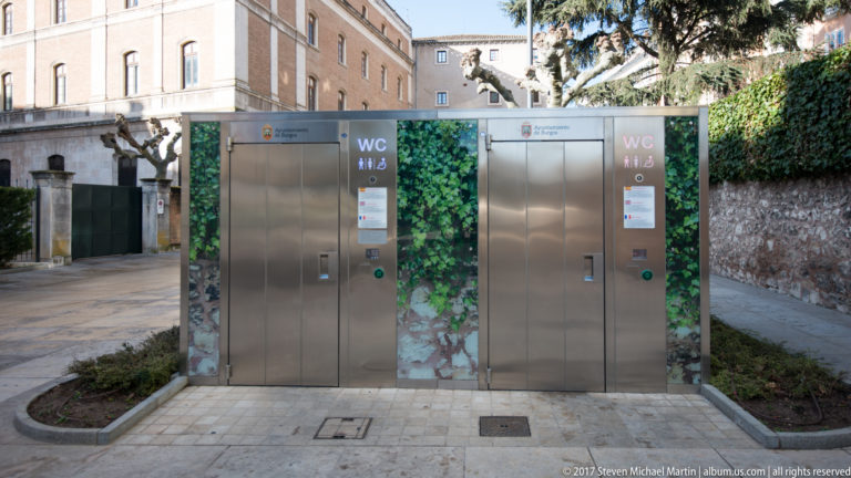Two stainless-steel public WC kiosks with leafy graphics beside a stone wall and landscaped path in Burgos.