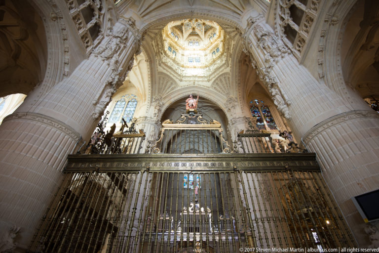 Tall iron grille enclosing an elevated monument beneath pale Gothic vaults and stained glass inside Burgos Cathedral.
