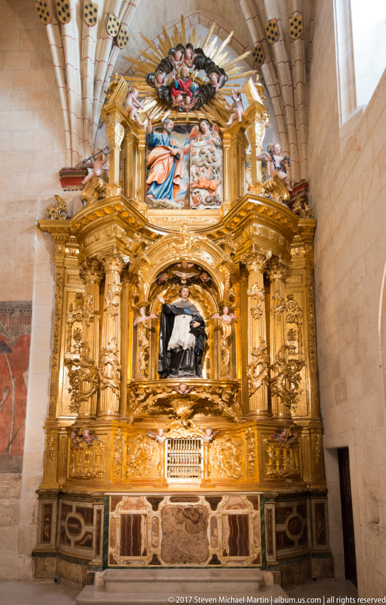 Large gilded wall monument with columns, carved ornament, painted figures, and a dark central niche inside Burgos Cathedral.