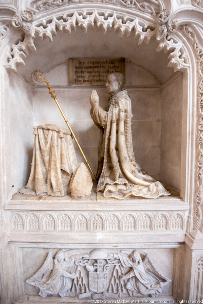 Pale stone kneeling effigy in long robes beside a draped prayer desk, set within an ornate carved wall niche.