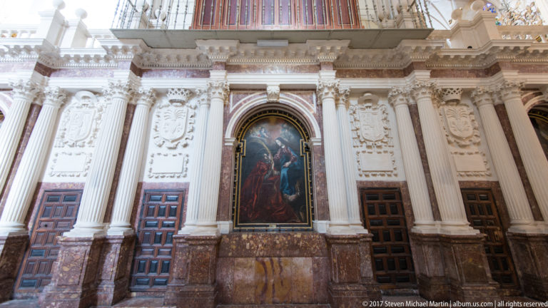 Tall white fluted columns and relief panels framing a religious painting above reddish marble and three dark wooden doors.