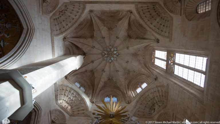 Upward view of intricate pale stone rib vault with central boss, high windows, and a gilded sunburst ornament.