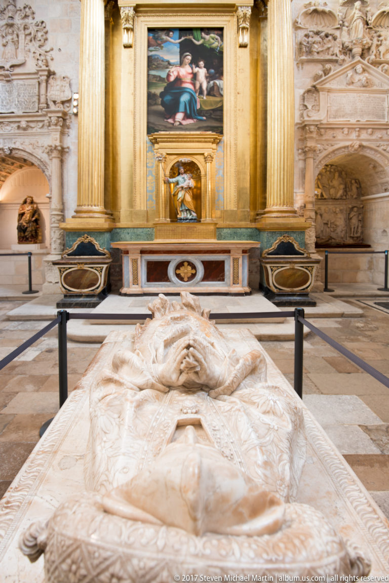 Recumbent pale stone tomb effigy in foreground with gilded-column altar and religious paintings behind.