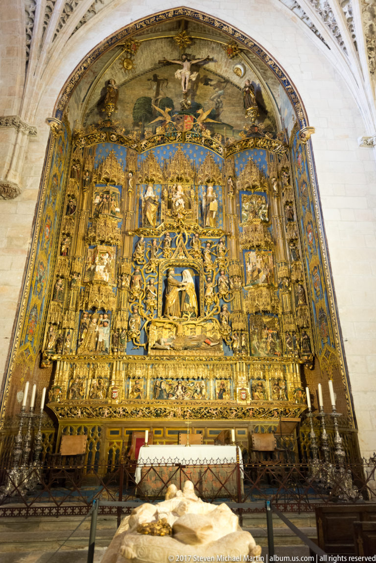 Large gilded and blue-accented retablo with many sculpted panels, above an altar and pale tomb effigy in foreground.