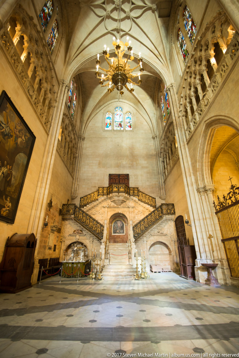 Wide interior view of Burgos Cathedral’s Golden Staircase with branching flights, gilded railings, central doorway, and ribbed vault.