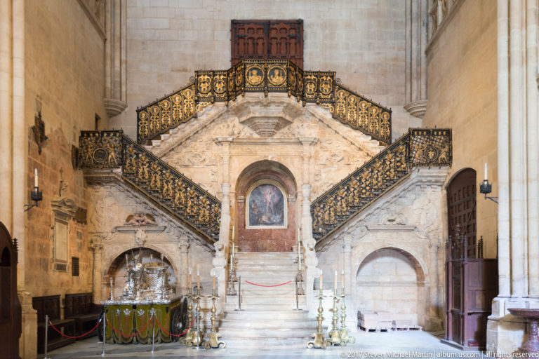 Frontal view of the Golden Staircase in Burgos Cathedral, with symmetrical stone flights and ornate black-and-gold railings.