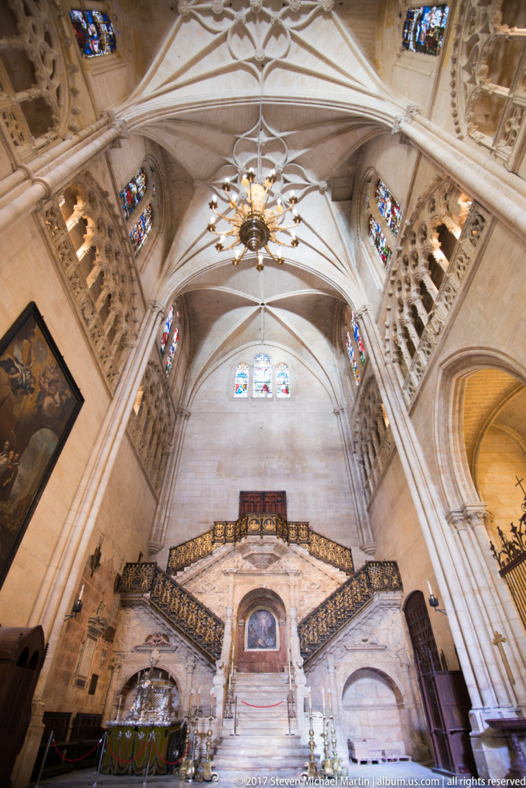 Wide vertical view of Burgos Cathedral’s Golden Staircase beneath ribbed vaults, stained glass, galleries, and chandelier.
