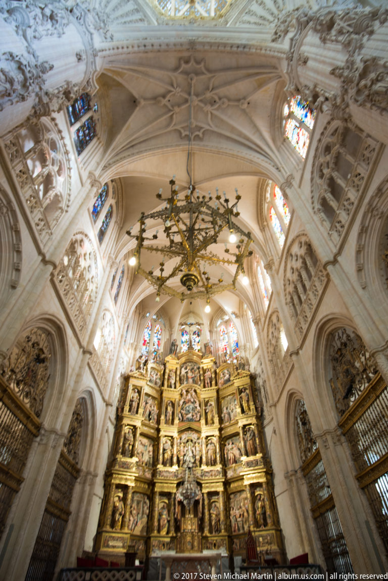 Tall gilded retablo beneath elaborate rib vaults, flanked by heraldic carving, stained glass, metal screens, and chandelier.