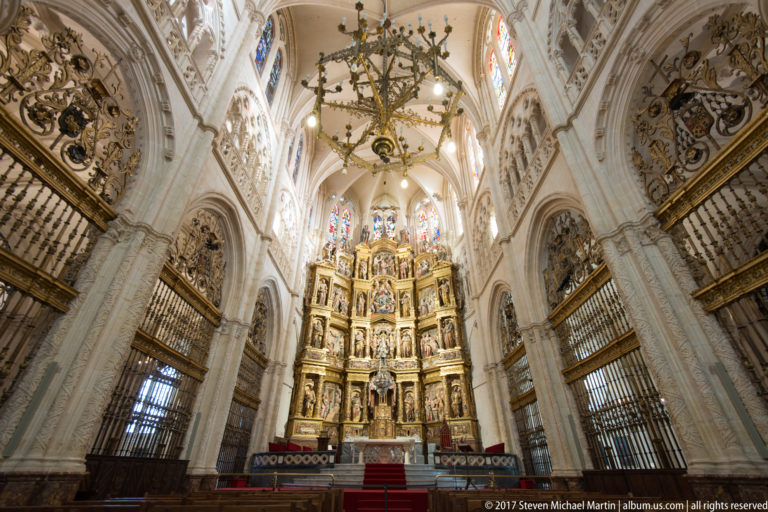 Frontal chapel interior with towering gilded retablo, rib vaults, stained glass, heraldic shields, and iron screens.