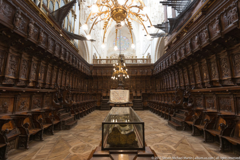 Long cathedral choir lined with dark carved wooden stalls, central lectern and open choir book beneath bright Gothic vaults.
