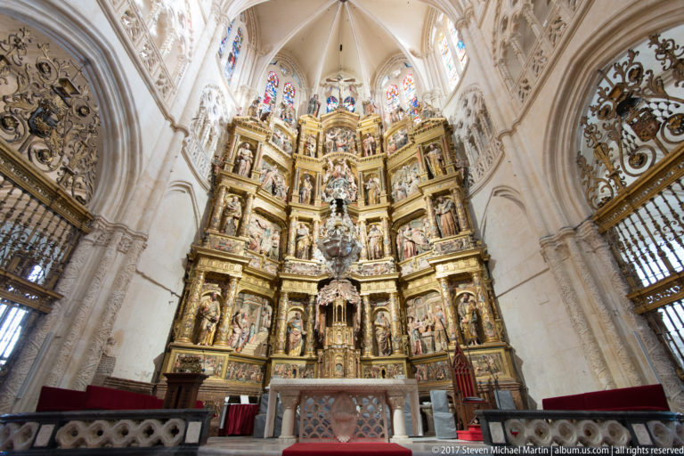 Close frontal view of towering gilded retablo with sculpted figures, narrative panels, altar table, red furnishings, and heraldic shields.